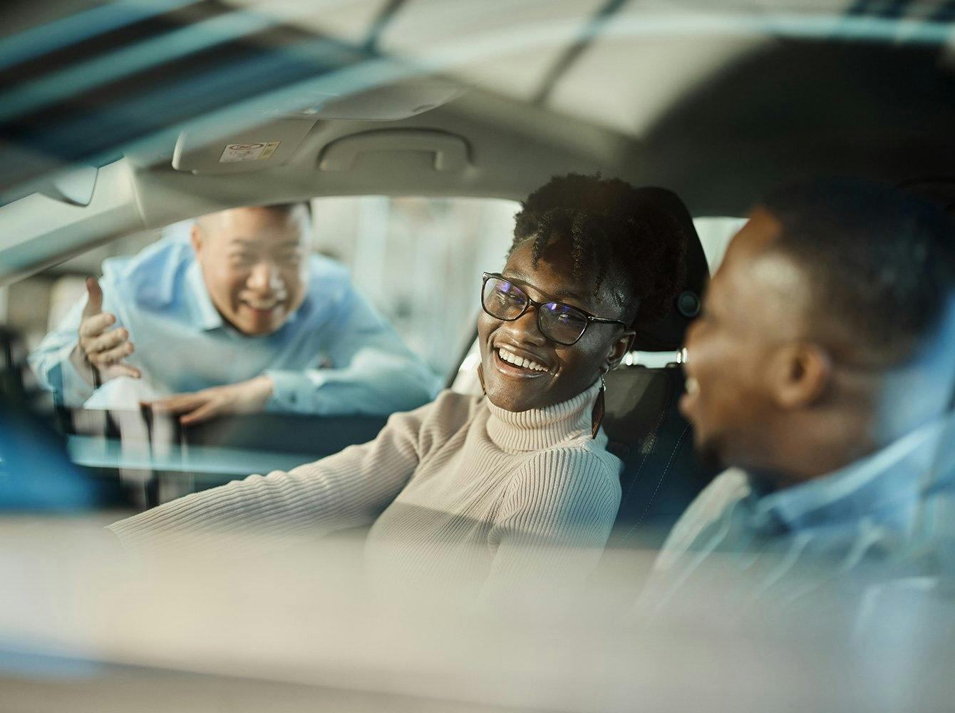 a smiling couple sitting in a car with a salesman talking to them at the window