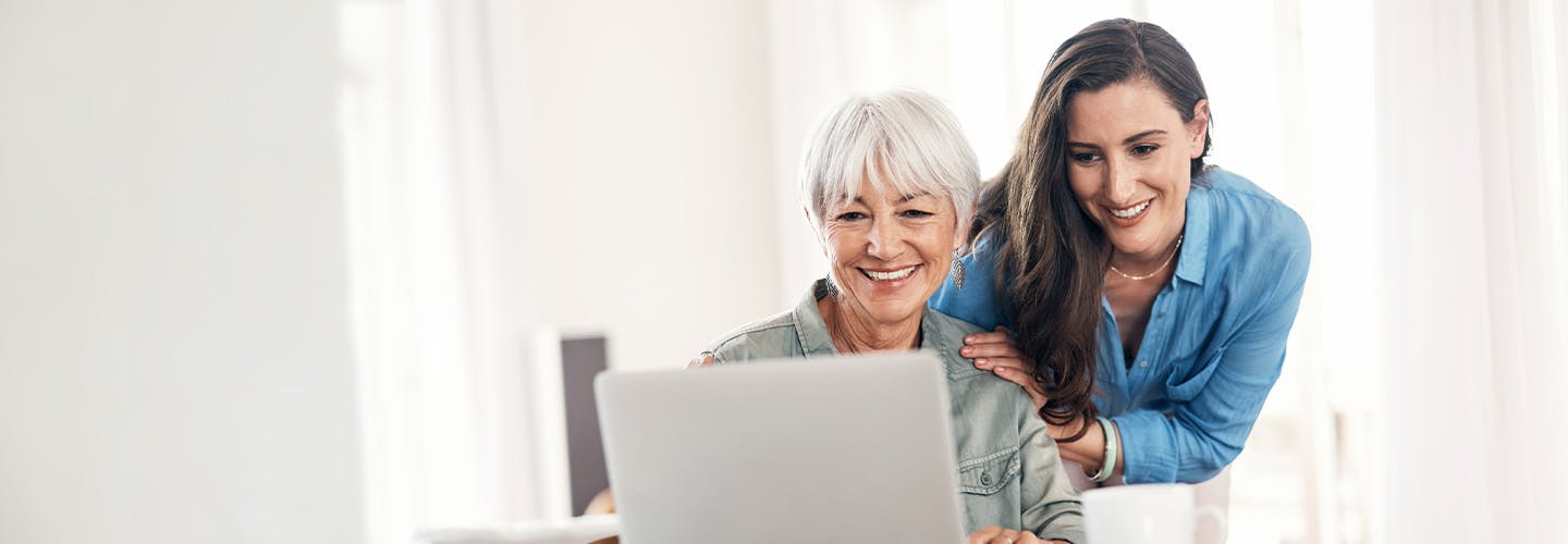 A younger woman stands behind an older woman helping her use a laptop.