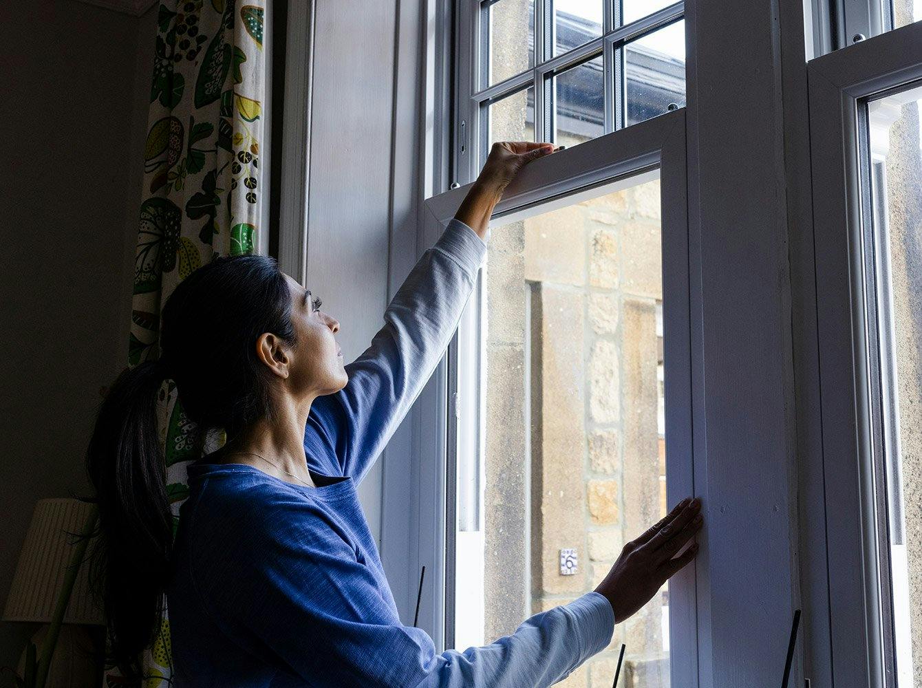 A woman reaching up to unlock a window in a house