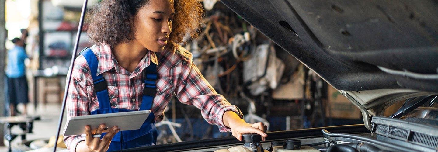 A woman with a tablet is working under the hood of a car