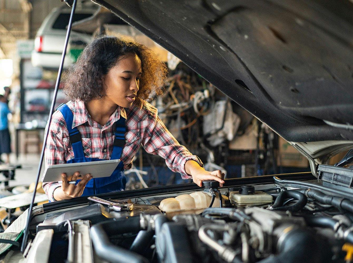 A woman with a tablet is working under the hood of a car